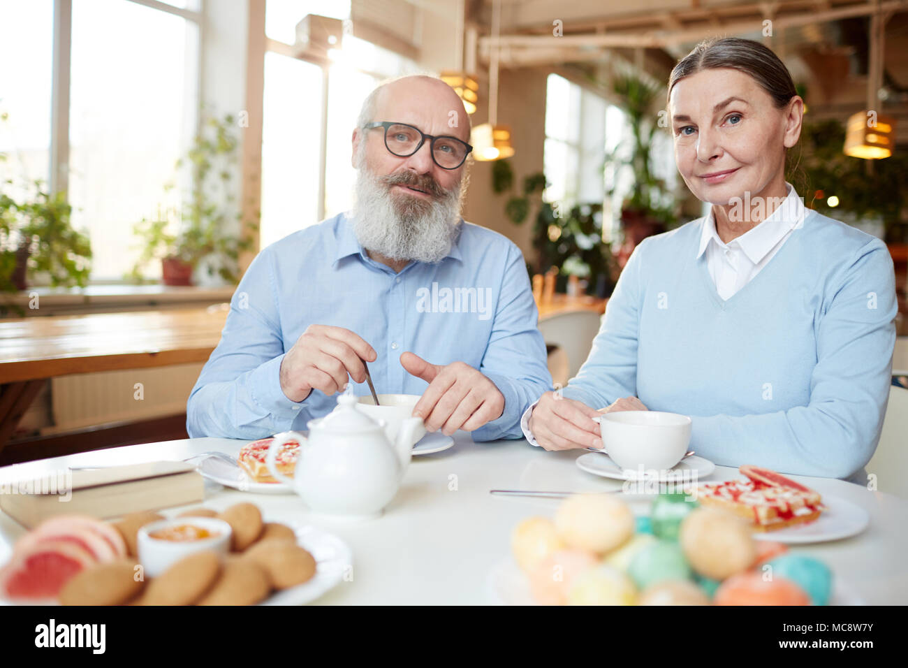 Senior couple sitting by table avec les cookies et desserts, boire du thé frais et looking at camera Banque D'Images