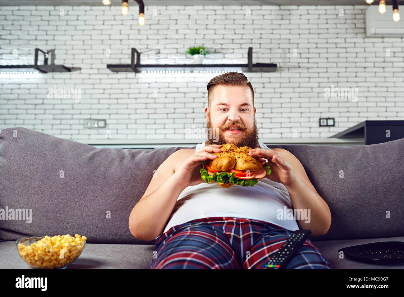 L'homme drôle Fat en pyjama de manger un hamburger assis sur le canapé à la  maison Photo Stock - Alamy