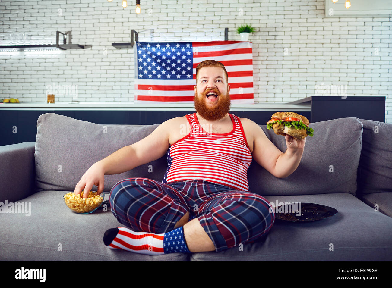 L'homme drôle épais de manger un burger assis sur le canapé, regarder la télévision à la maison contre le fond du drapeau américain. Banque D'Images