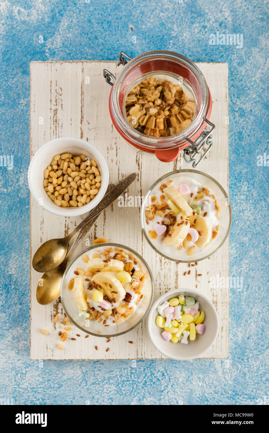 Healthy dessert de yaourt, banane, granola et les écrous dans verre verres sur une planche à découper en bois blanc sur fond bleu. Vue d'en haut Banque D'Images