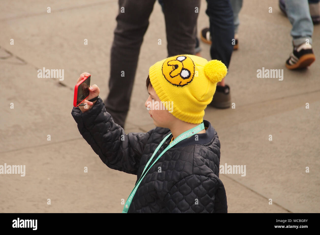 Un jeune garçon tenant un sur son smartphone selfies à Trafalgar Square, Londres, portant un chapeau jaune bobble et matelassée manteau hivers Banque D'Images