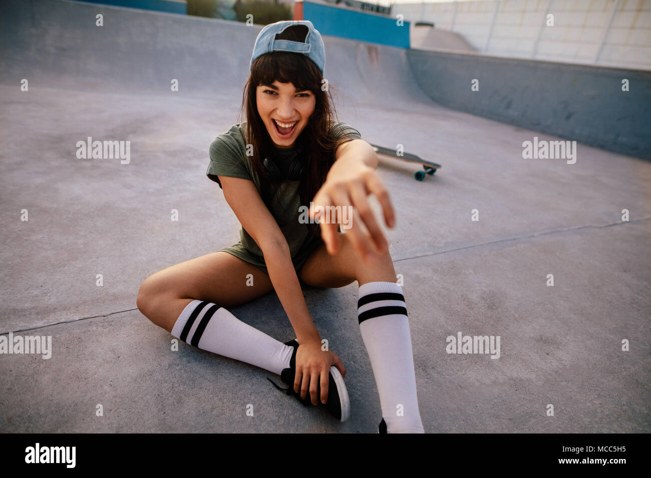 Laughing young skateboarder sitting at skate park et en montrant l'appareil photo. Cheerful woman outdoors at skate park s'amusant. Banque D'Images