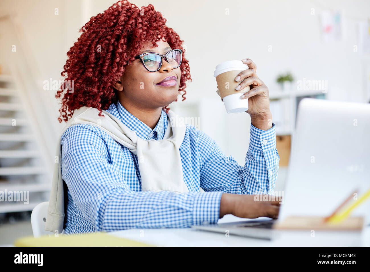 Portrait of attractive African American Woman with red cheveux bouclés assis à un ordinateur portable avec du café à emporter et sourire rêveur Banque D'Images