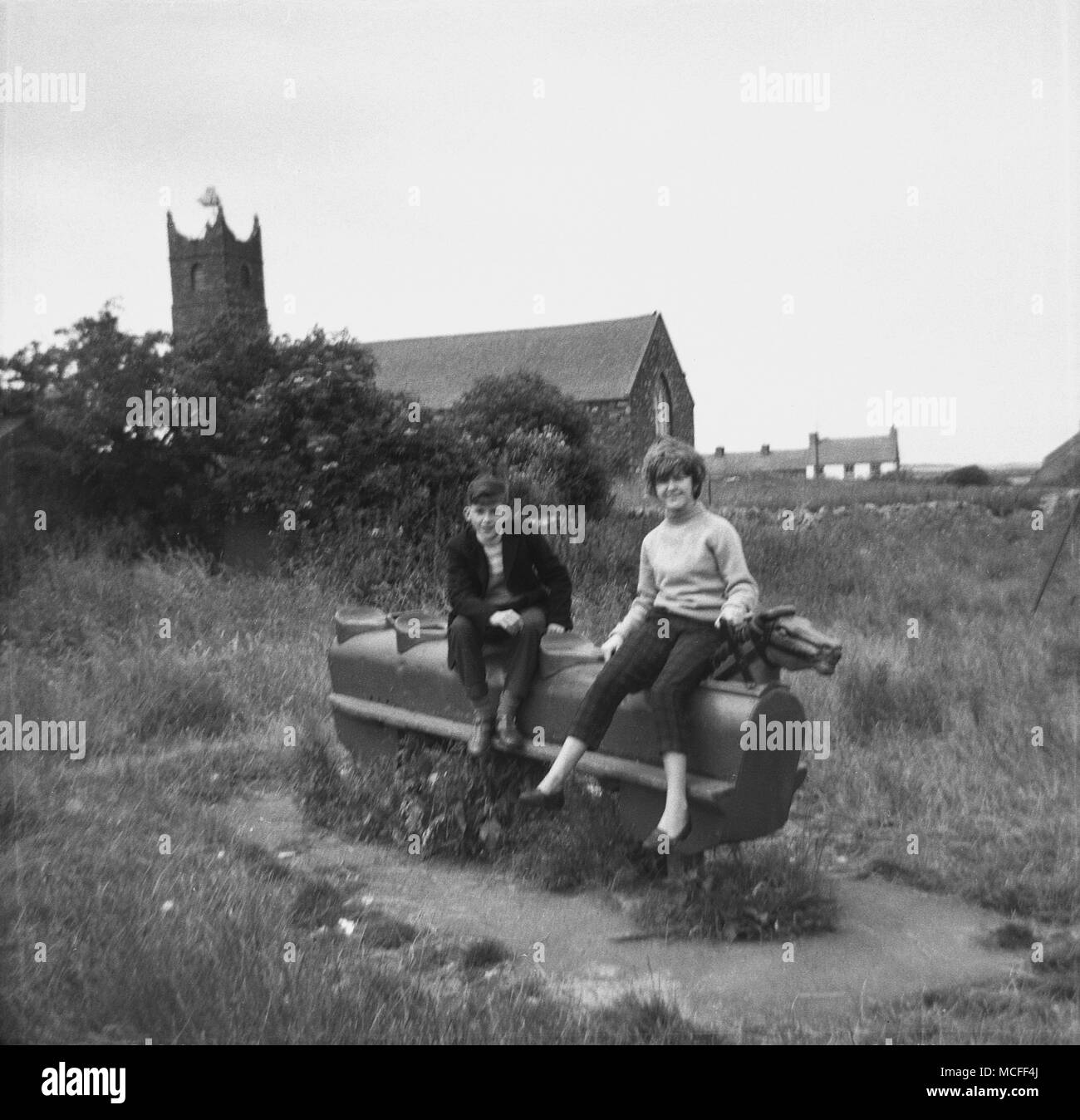 Liverpool, UK,1960, après l'école, deux adolescents, un garçon et une fille, assise à l'extérieur sur un vieux cheval à bascule en métal à une aire de jeux herbeuse derrière une église. Banque D'Images