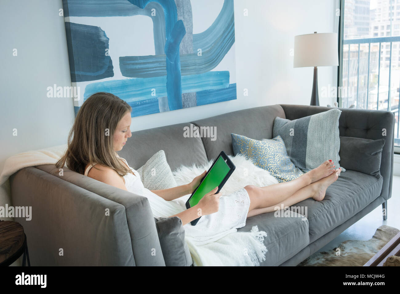 Young woman using digital tablet while lying on sofa in living room Banque D'Images