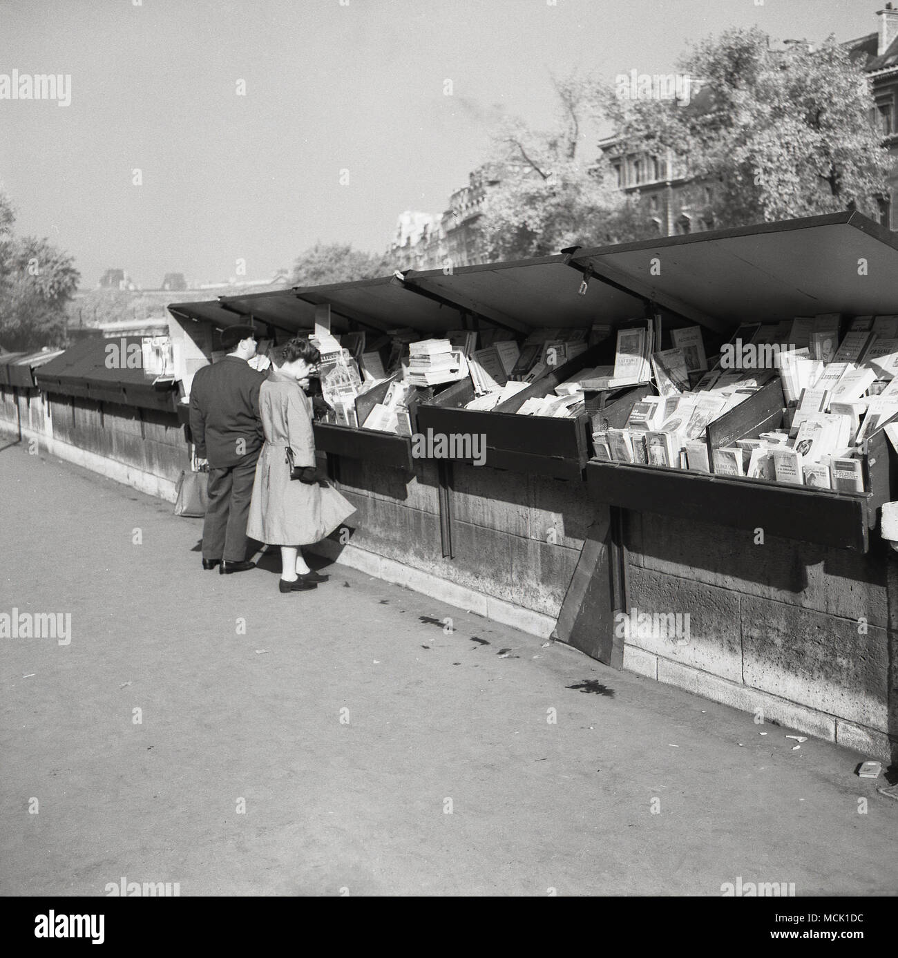 Années 1950, tableau historique d'un couple parisien le stock à un bouquiniste à Paris, France. Ces libraires indépendants de utilisé et le commerce de livres anciens à partir de boîtes de métal le long des sections de la rives de la Seine et ont été une caractéristique importante de la vie parisienne depuis plus de 100 ans. Banque D'Images