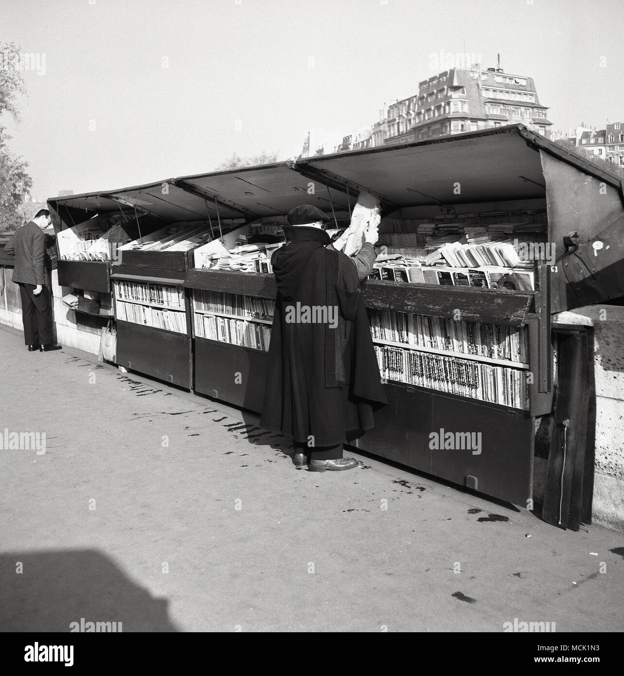 Années 1950, tableau historique d'un homme portant un béret Parisien et une longue cape en regardant le stock à un bouquiniste à Paris, France. Ces libraires indépendants de utilisé et le commerce de livres anciens à partir de boîtes de métal le long des sections de la rives de la Seine et ont été une caractéristique importante de la vie parisienne depuis plus de 100 ans. Banque D'Images
