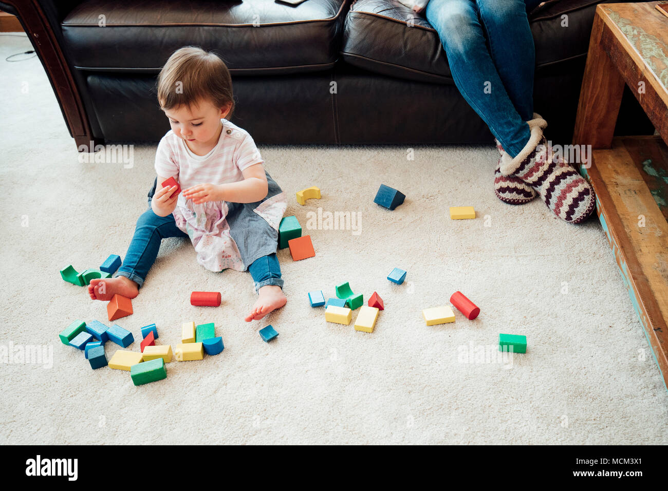 Petite fille est assise sur le tapis dans le salon à jouer avec des blocs de jouets. Sa mère est assise sur le canapé derrière elle. Banque D'Images