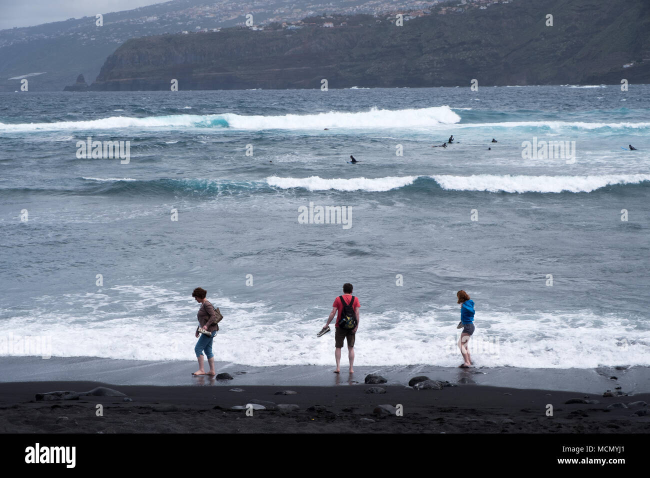 Puerto de la Cruz, Tenerife, Canaries ; touristes tester la température de l'eau sur une plage de sable noir alors que les surfeurs pièges pour prendre les vagues. Banque D'Images