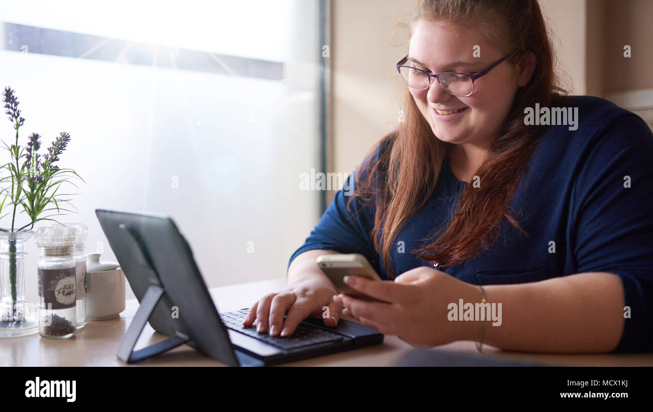 Caucasian girl smiling en regardant son téléphone portable pendant son cappachino du matin alors qu'elle consulte ses mails sur une tablette électronique dans son café préféré avec une lumière vive. Banque D'Images