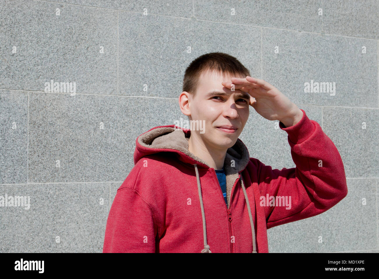 Le gars dans la capuche rouge et se tient au mur et couvre ses yeux avec sa main de la lumière du soleil. Banque D'Images