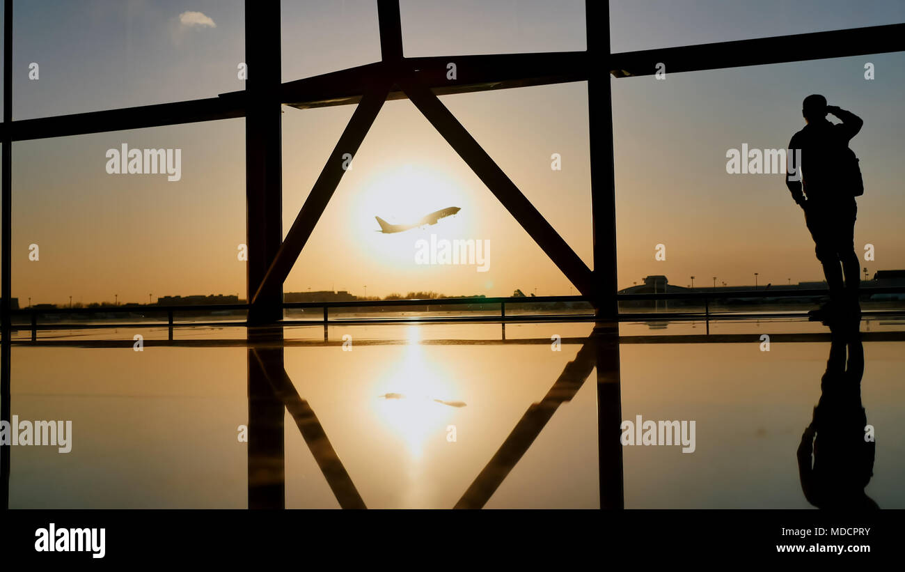 Silhouette d'un tourist guy regarder le décollage de l'avion debout à la fenêtre de l'aéroport au coucher du soleil le soir. Concept de voyage, les gens de l'aéroport. Banque D'Images