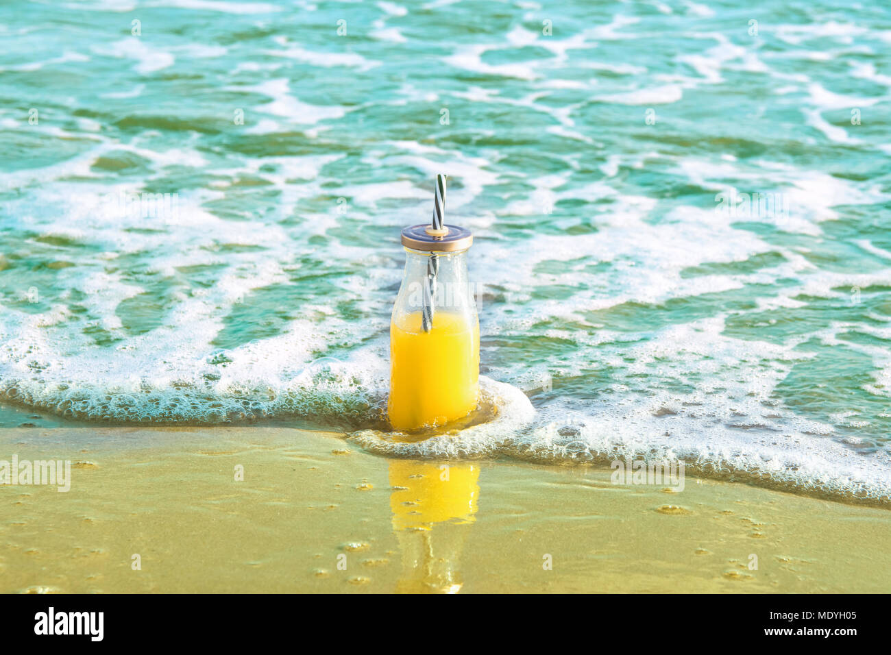 Le flacon en verre avec des Jus Jus de fruits tropicaux avec de la paille dans des éclaboussures sur le sable de la plage baignée par le bleu turquoise de la mer des vagues. mousseux Golden Banque D'Images