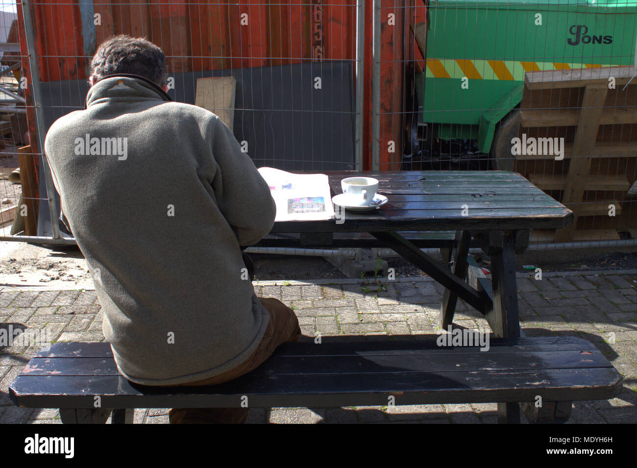 L'homme de prendre une pause café le matin et lire son journal sur un banc en bois et une table à l'extérieur d'un café, dans le soleil du matin. Banque D'Images