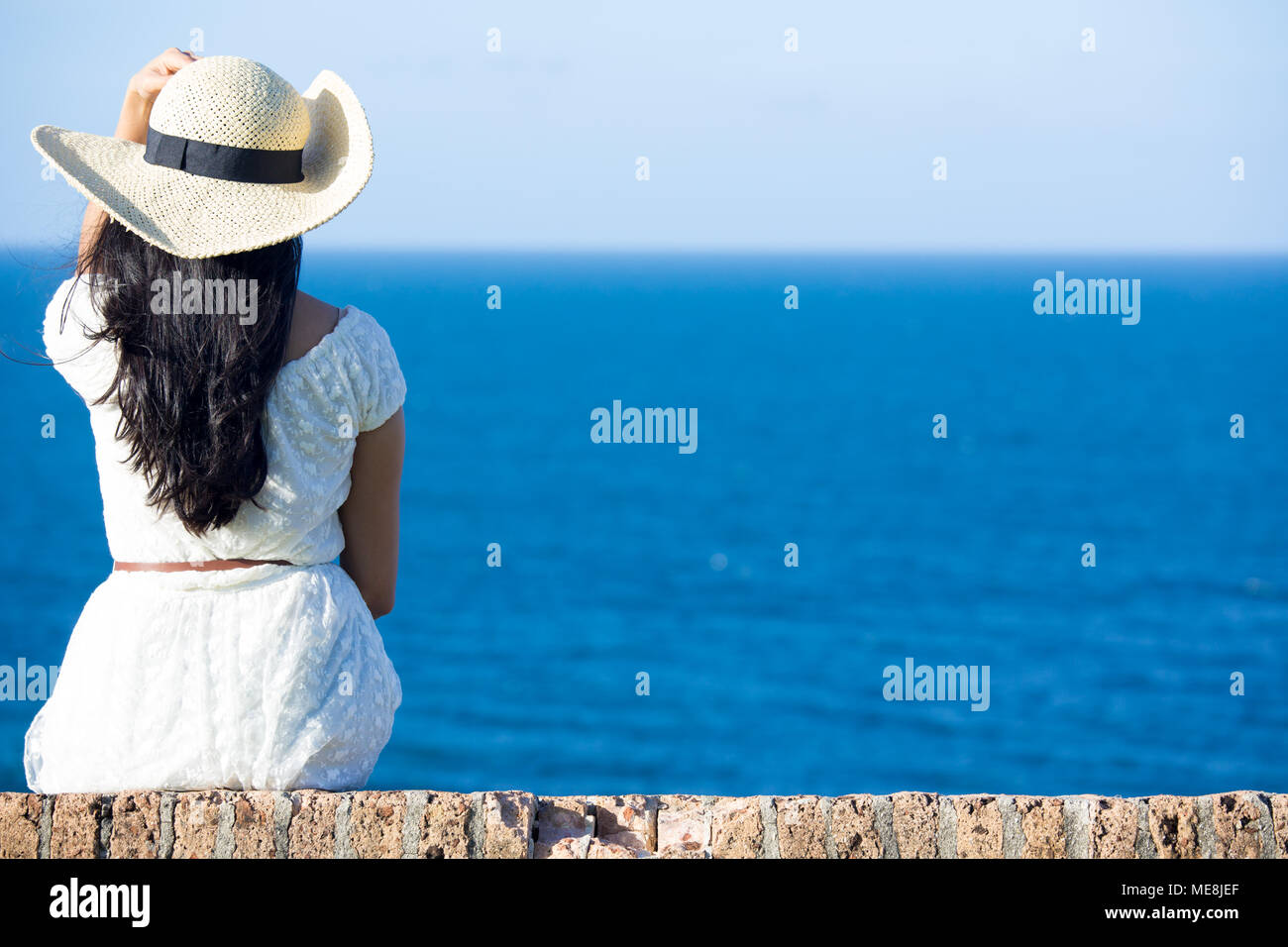 Vue arrière de gros plan femme assise en robe blanche et un chapeau à l'extérieur, vers l'océan et le ciel bleu, l'arrière-plan la mer isolés Banque D'Images