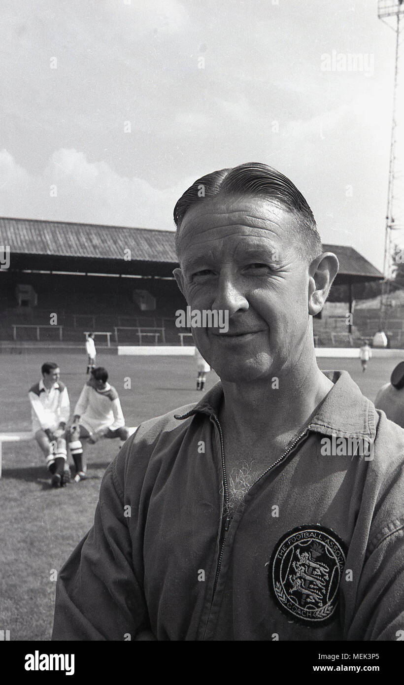 1964, tableau historique, à l'entraîneur de Charlton Athletic FC à leur terrain de football, la vallée, avec des joueurs derrière lui sur le terrain. Sa partie supérieure comporte une grande d'un insigne indiquant qu'il est un entraîneur de l'English Football Association (FA) Banque D'Images
