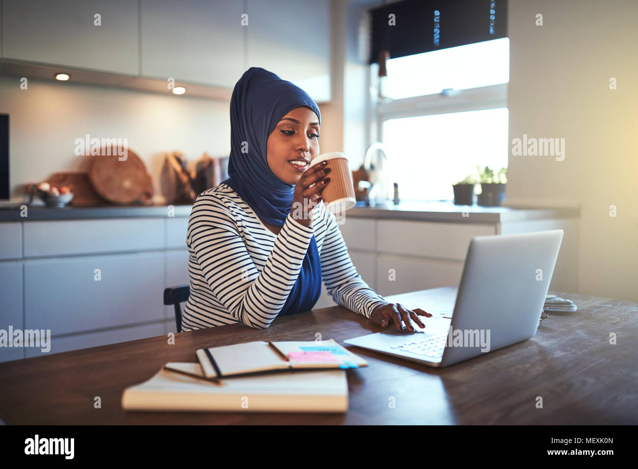Jeune femme entrepreneur arabe portant un hijab de boire un café et de travailler sur un ordinateur portable tout en étant assis à une table dans sa cuisine Banque D'Images