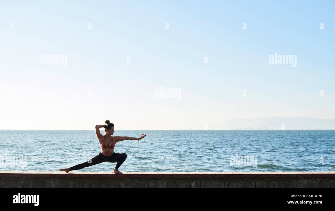 Young woman practicing yoga sur un mur par la mer Banque D'Images