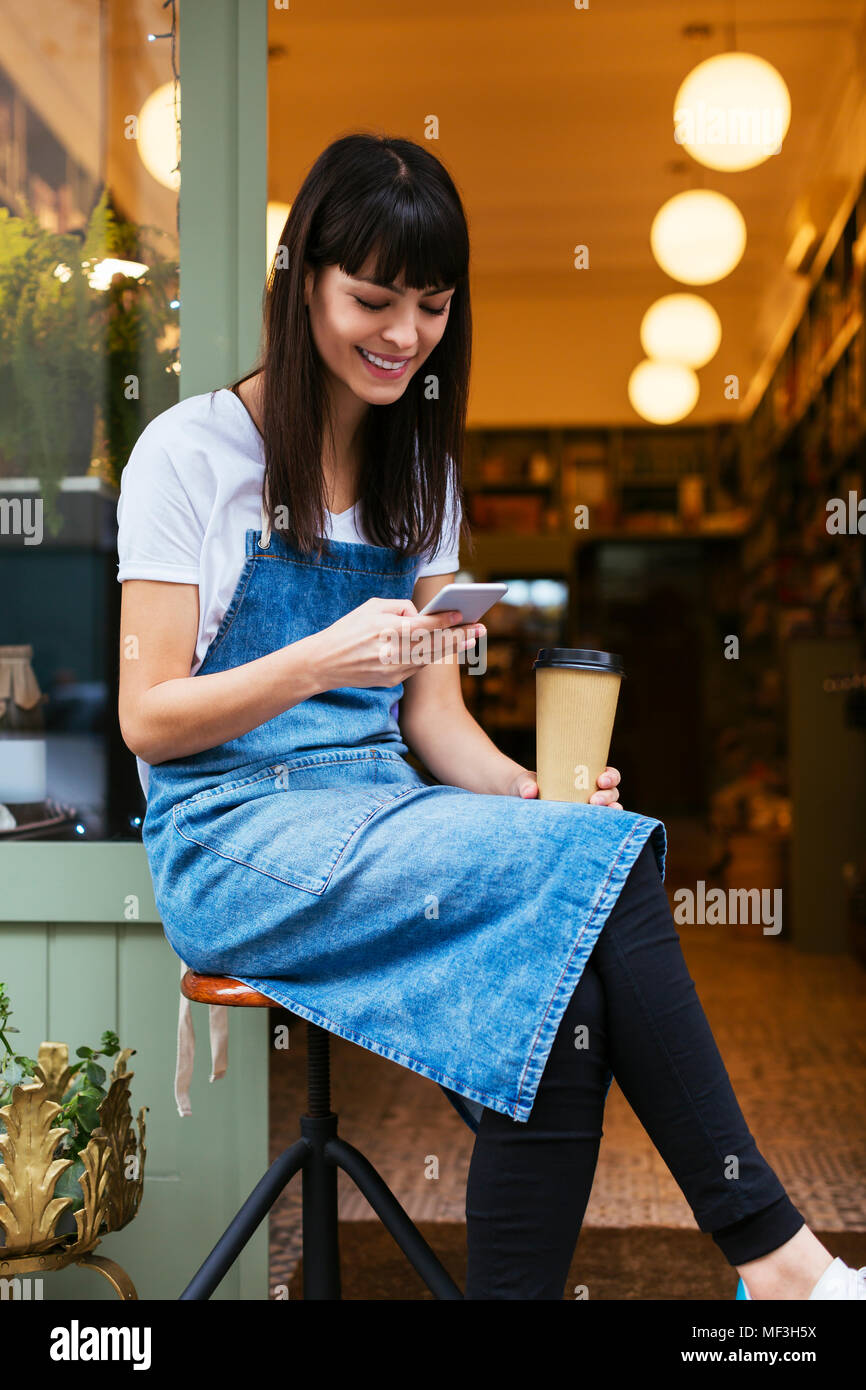 Smiling woman sitting on stool using cell phone at porte d'entrée d'un magasin Banque D'Images