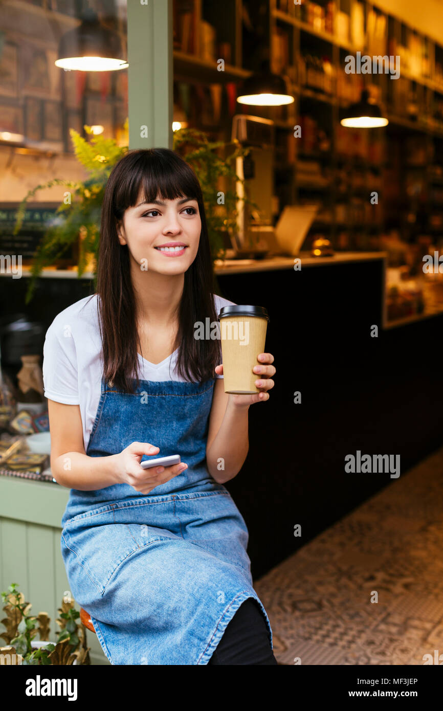 Smiling woman sitting at porte d'entrée d'un magasin holding cell phone et café à emporter Banque D'Images