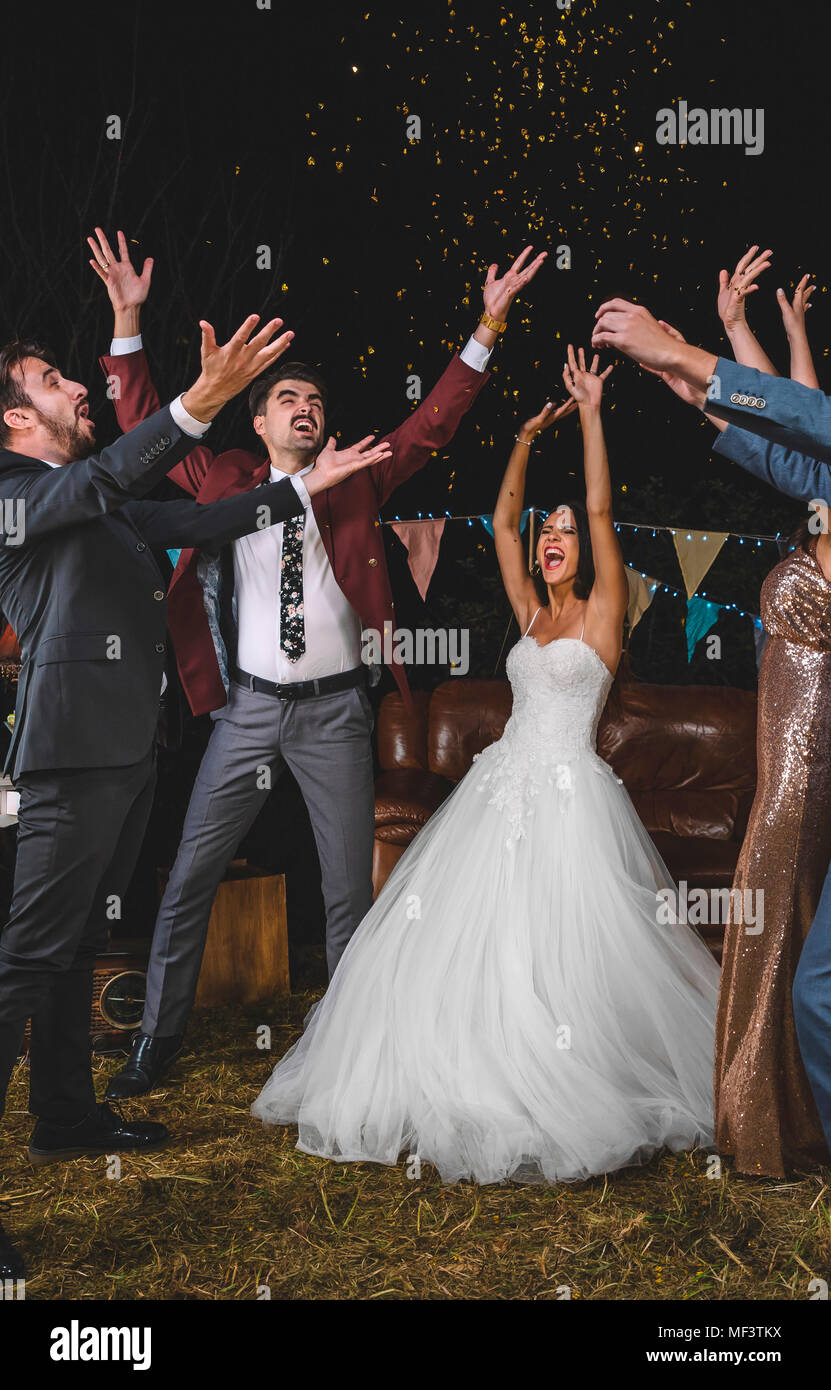 Couple de mariage gai et amis levant les bras tandis que les confettis tombant sur la tête d'un parti à l'extérieur de nuit Banque D'Images