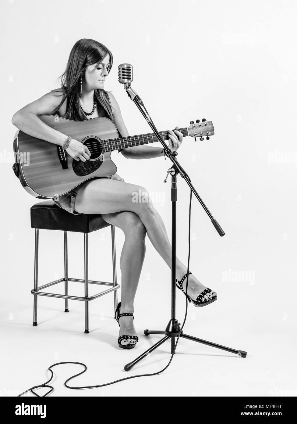 Photo d'une jeune femme aux cheveux longs assis sur un tabouret pour jouer une guitare acoustique sur fond blanc. Banque D'Images