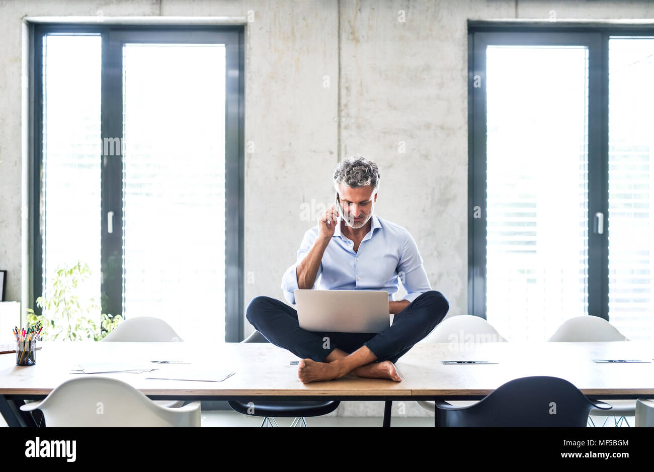 Mature businessman sitting on desk in office pieds nus à l'aide d'un téléphone cellulaire et ordinateur portable Banque D'Images