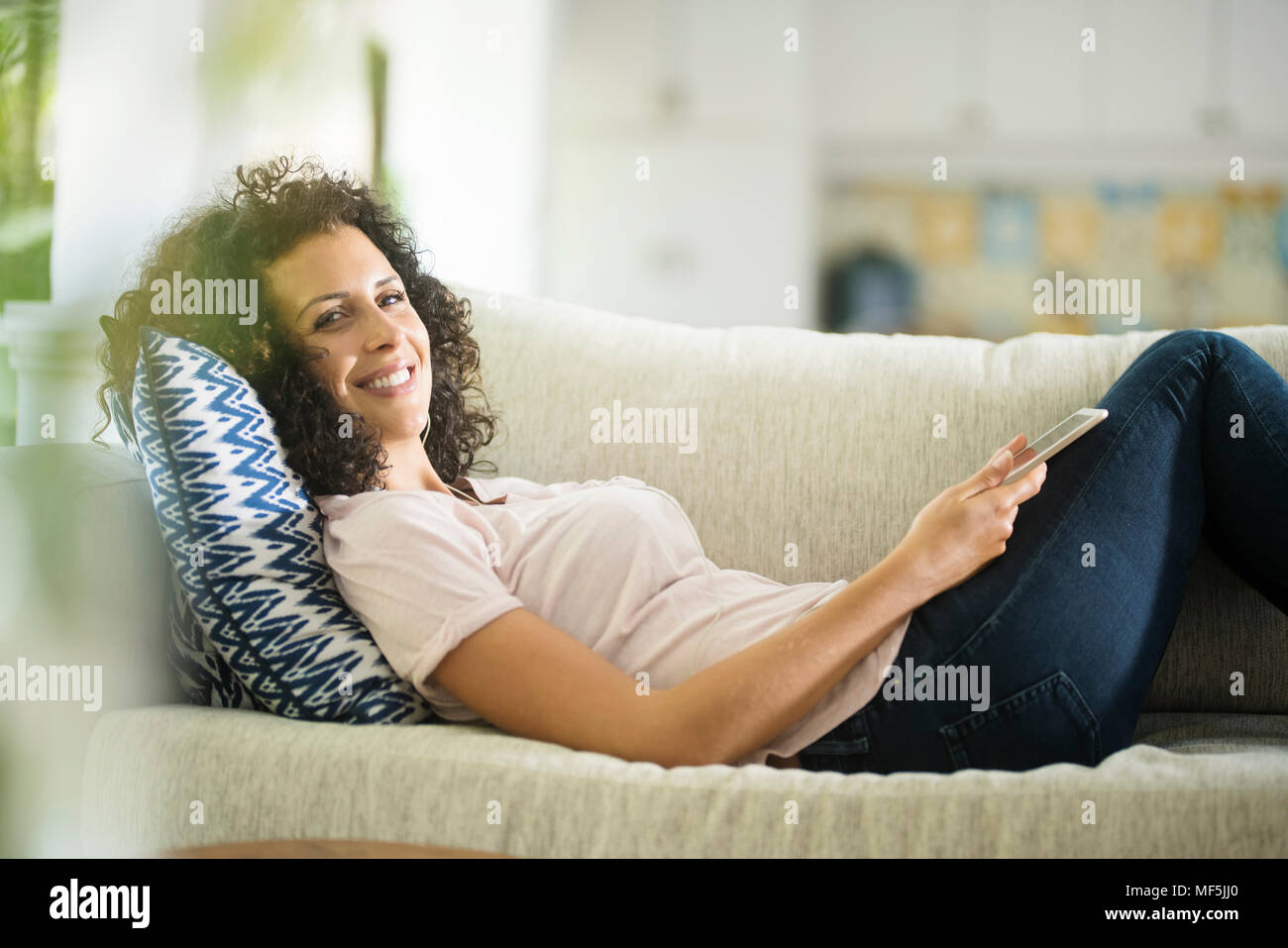 Femme souriante avec des cheveux bouclés lying on sofa holding tablet Banque D'Images