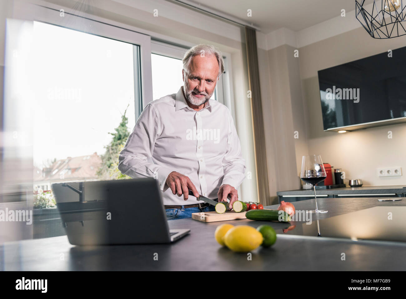 Homme mûr de préparer des aliments dans la cuisine while looking at laptop Banque D'Images