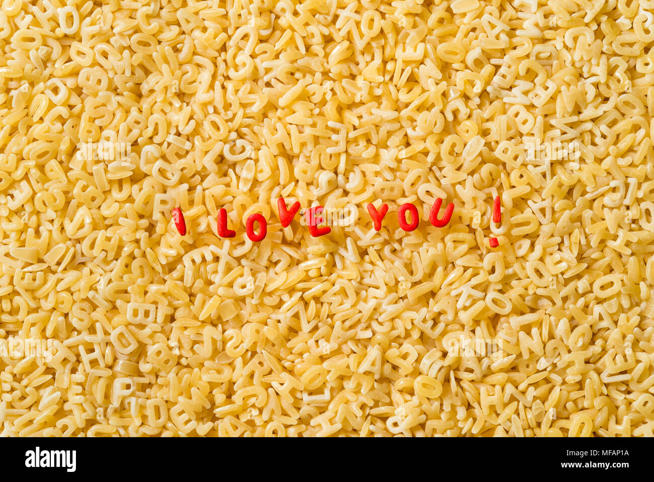 Les mots « Je t'aime ! » écrits avec des lettres de pâtes abc de couleur rouge. Le fond est fait de lettres de pâtes abc naturelles non cuites. Banque D'Images