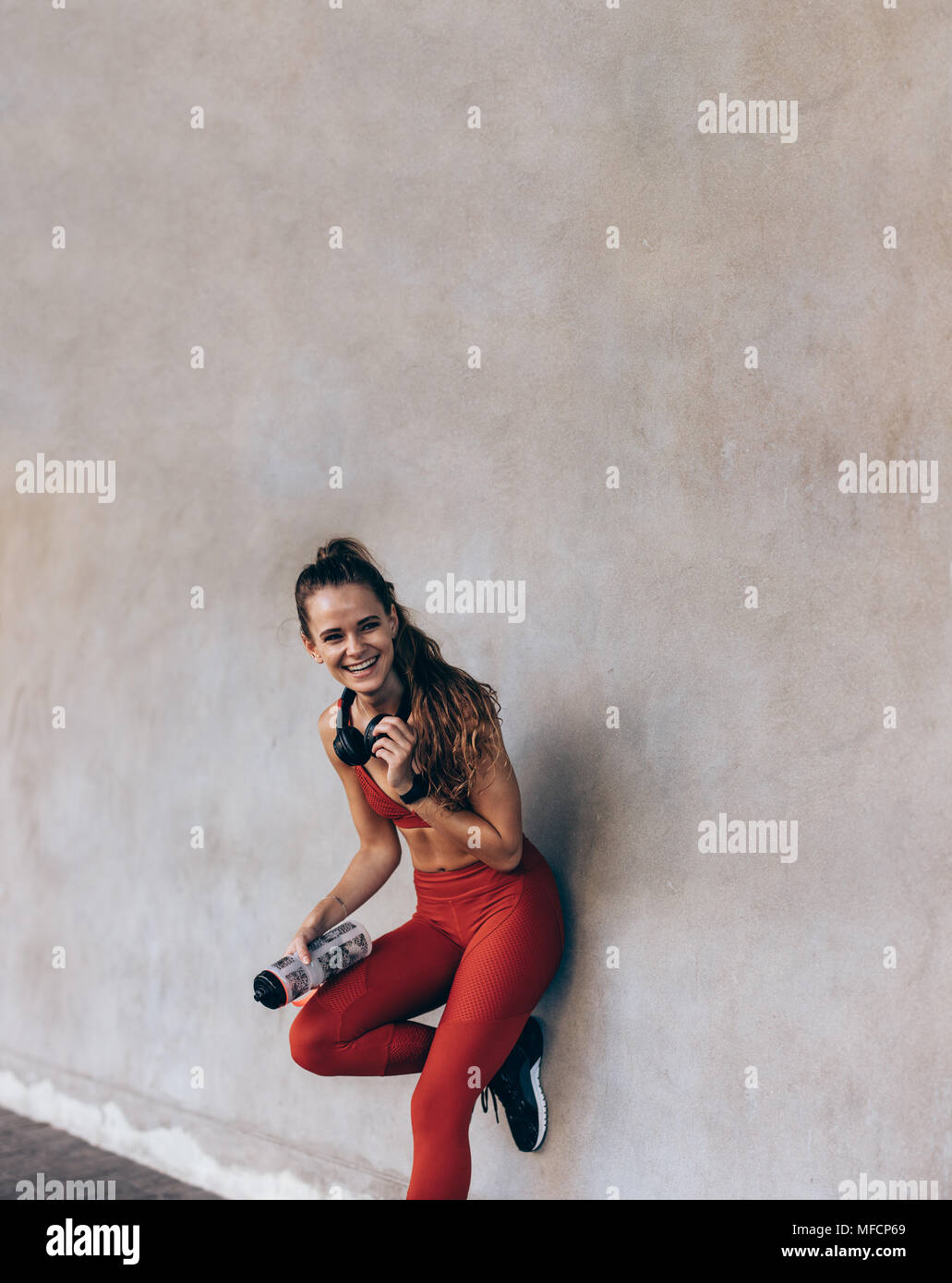 Souriante jeune femme sportswear comité permanent par un mur gris. Athlète féminin avec de l'eau en bouteille après pause session de formation à l'extérieur. Banque D'Images