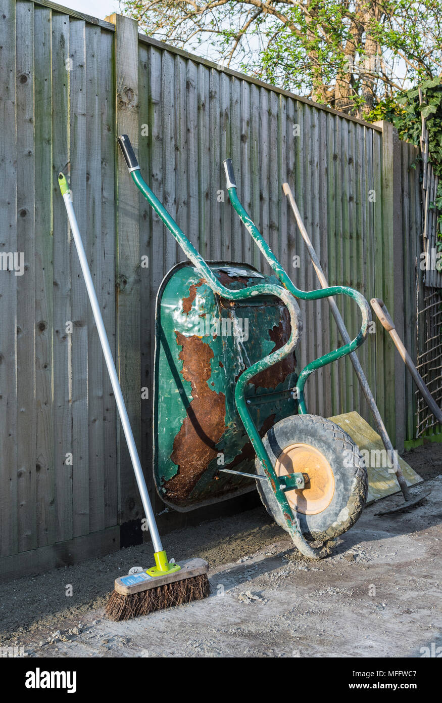 Brouette et autres outils de jardin s'appuyant sur une clôture à la fin de la journée de travail pendant les travaux de construction dans un jardin ou cour arrière. Banque D'Images