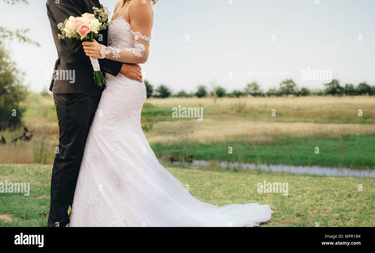 Debout à côté de la mariée le marié dans un parc. Femme dans une longue robe blanche avec un bouquet de fleurs dans ses mains. Cropped shot with copy space. Banque D'Images Debout à côté de la mariée le marié dans un parc. Femme dans une longue robe blanche avec un bouquet de fleurs dans ses mains. Cropped shot with copy space. Banque D'Images