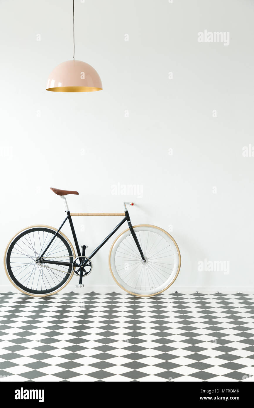 Vélo noir contre mur blanc en intérieur simple avec sol en damier et lampe rose Banque D'Images