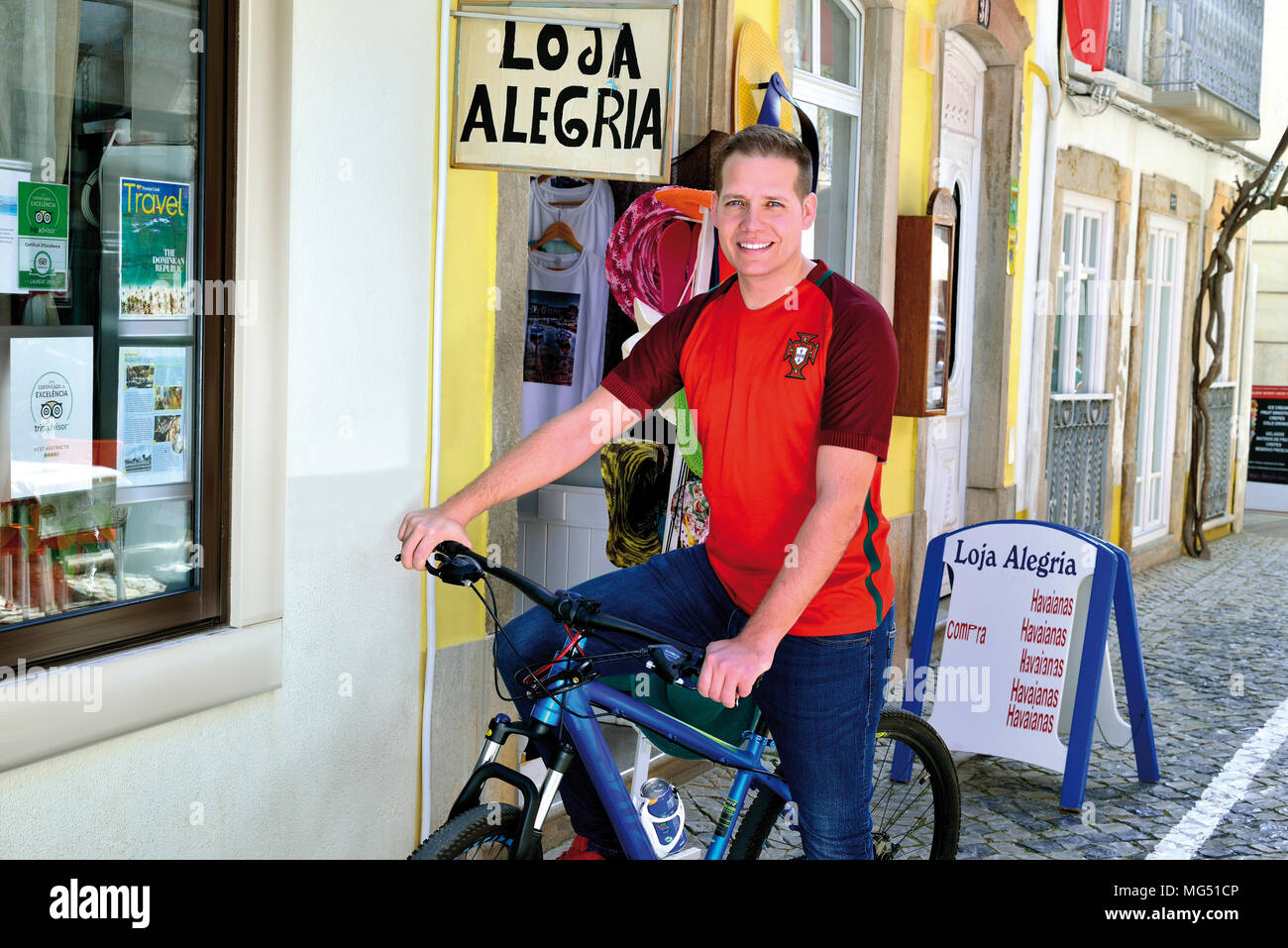 Jeune homme blond avec maillot de l'équipe nationale de football portugais assis sur un vélo et souriant à l'appareil photo Banque D'Images