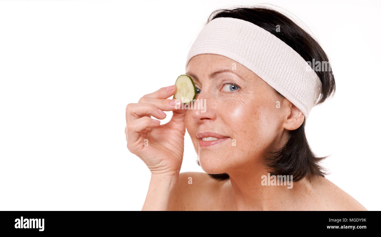 Cheerful mature woman holding morceau de concombre à l'avant de l'œil. Traitement spa naturel. Isolé sur fond blanc Banque D'Images