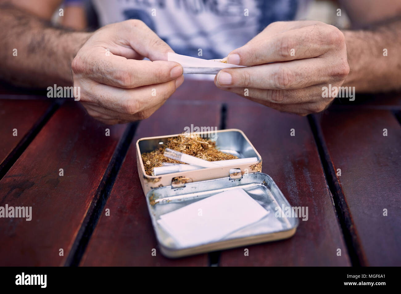 Les mains d'un homme de race blanche rouler une cigarette sur une table en bois. Papier à cigarettes, filtres, et une jolie boîte pleine de tabac est sur la table. Banque D'Images