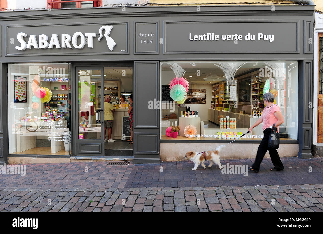 Une femme entre son chien en face de la boutique gastronomique Sabarot Each Qty au Puy-en-Velais, France Banque D'Images
