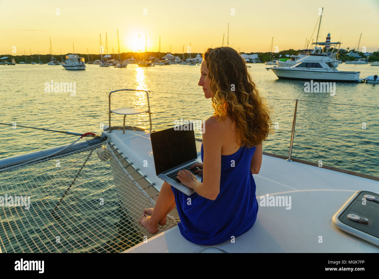 Jeune Femme au chapeau de soleil et lunettes travaille sur un ordinateur portable à la proue d'un voilier, bateaux et phare en arrière-plan Banque D'Images