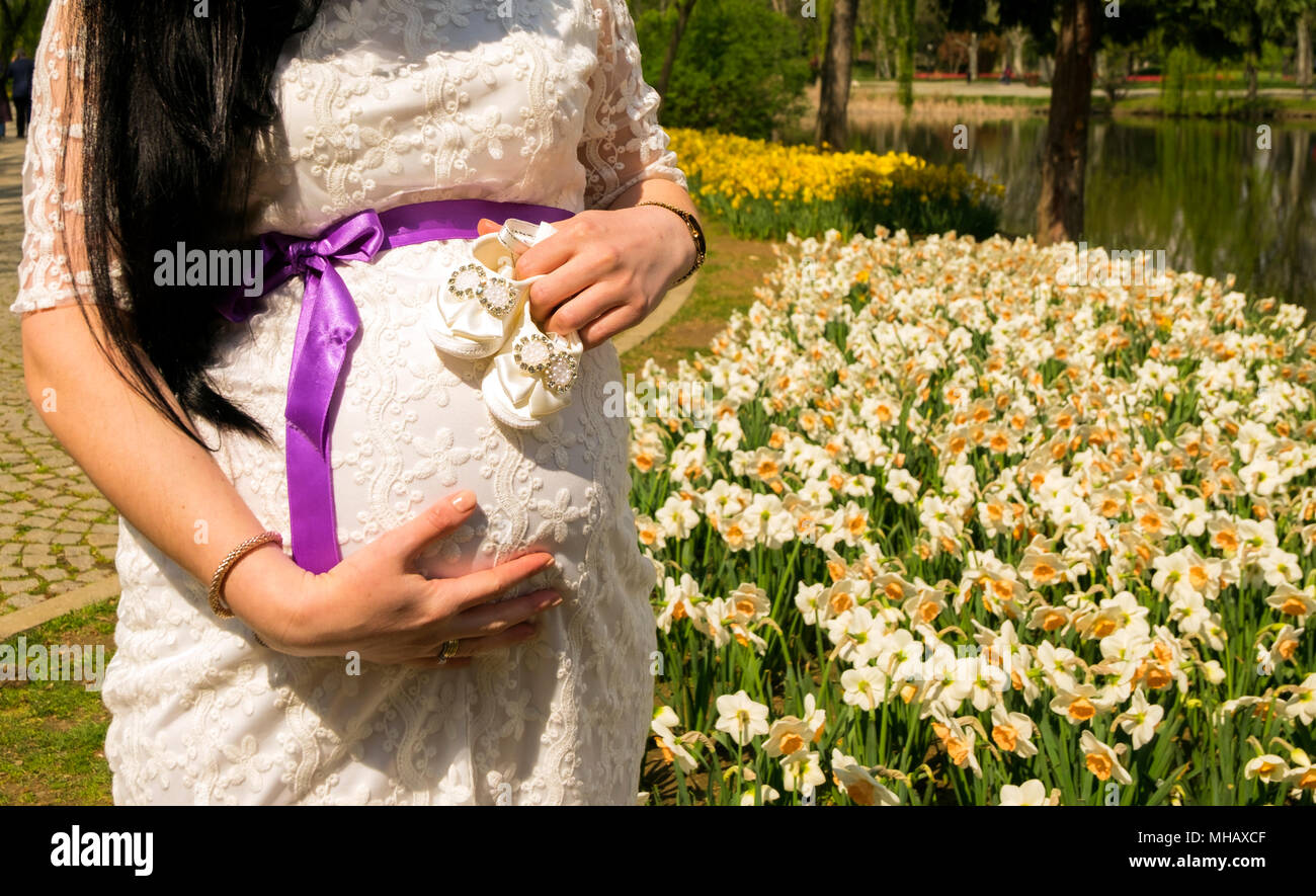 La femme enceinte attend son bébé. Cette photographie prend alors qu'elle tenait des chaussures dans sa main devant des fleurs et un lac. Banque D'Images