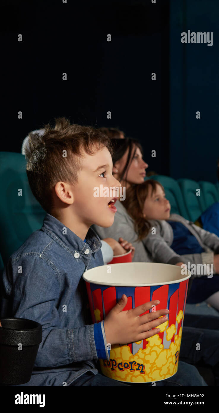 Cute boy avec la bouche ouverte et ferme les yeux en regardant le film dans un cinéma moderne. Petit viewer holding popcorn sac de papier. Petite fille assise sur sa mère tours et profiter de film en arrière-plan. Banque D'Images