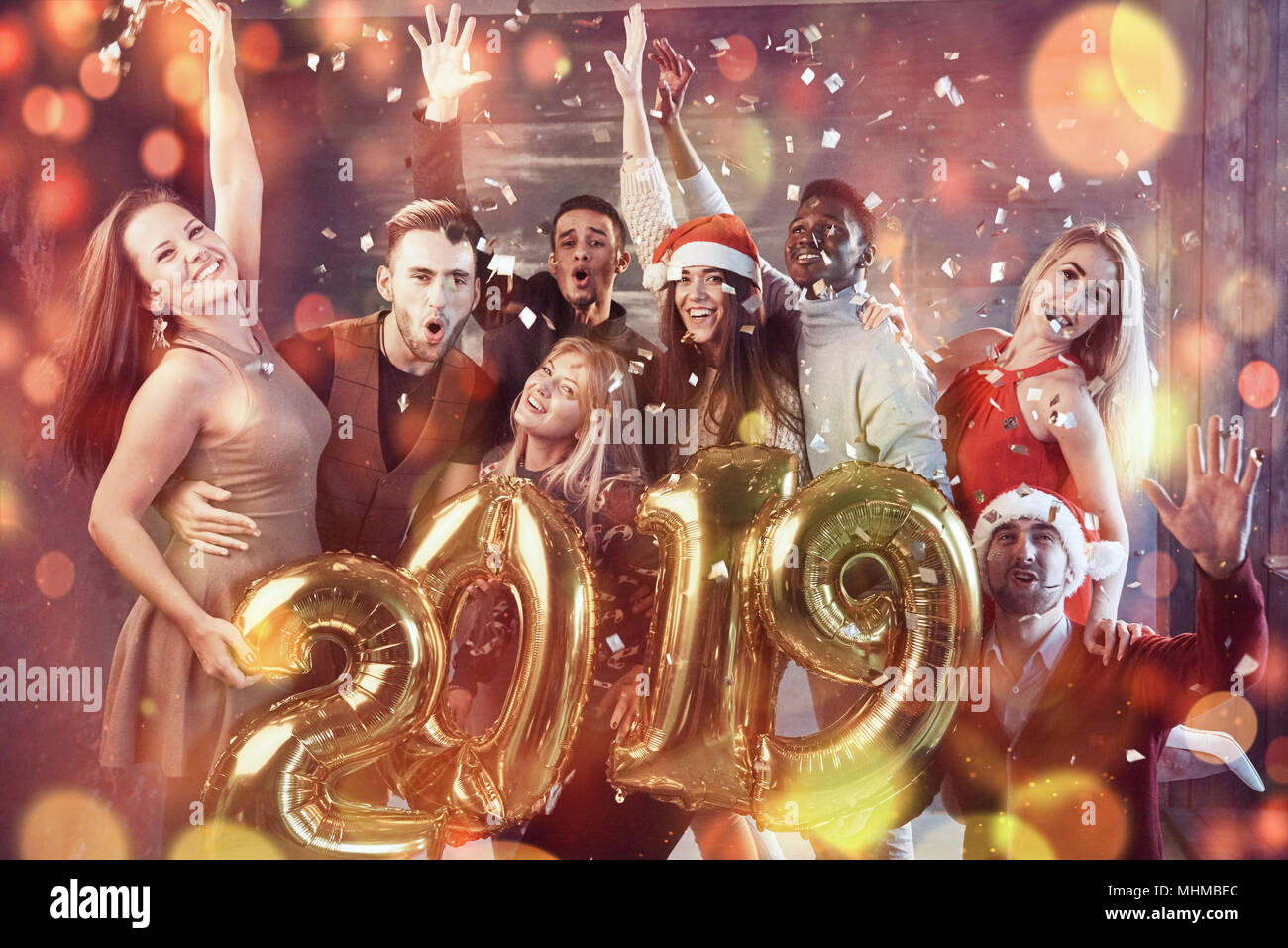 2019 Nouvelle année approche groupe de jeunes gens joyeux multiethnique dans Santa hats transportant l'or nombres en couleur et en jetant des confettis sur la partie Banque D'Images