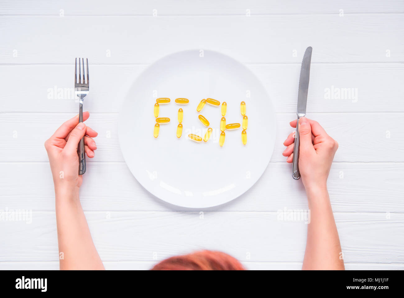 Vue d'en haut les mains tenant le couteau et la fourchette sous plaque ronde avec lettrage mot de poisson par l'huile de foie de morue Capsules Omega 3, sur la table en bois blanc. Banque D'Images