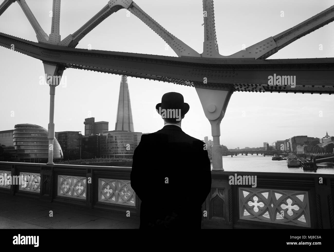 Vue arrière d'un homme portant manteau noir et chapeau melon debout sur le Tower Bridge sur la Tamise à Londres, en Angleterre, l'Hôtel de Ville et le Shard skyscrap Banque D'Images