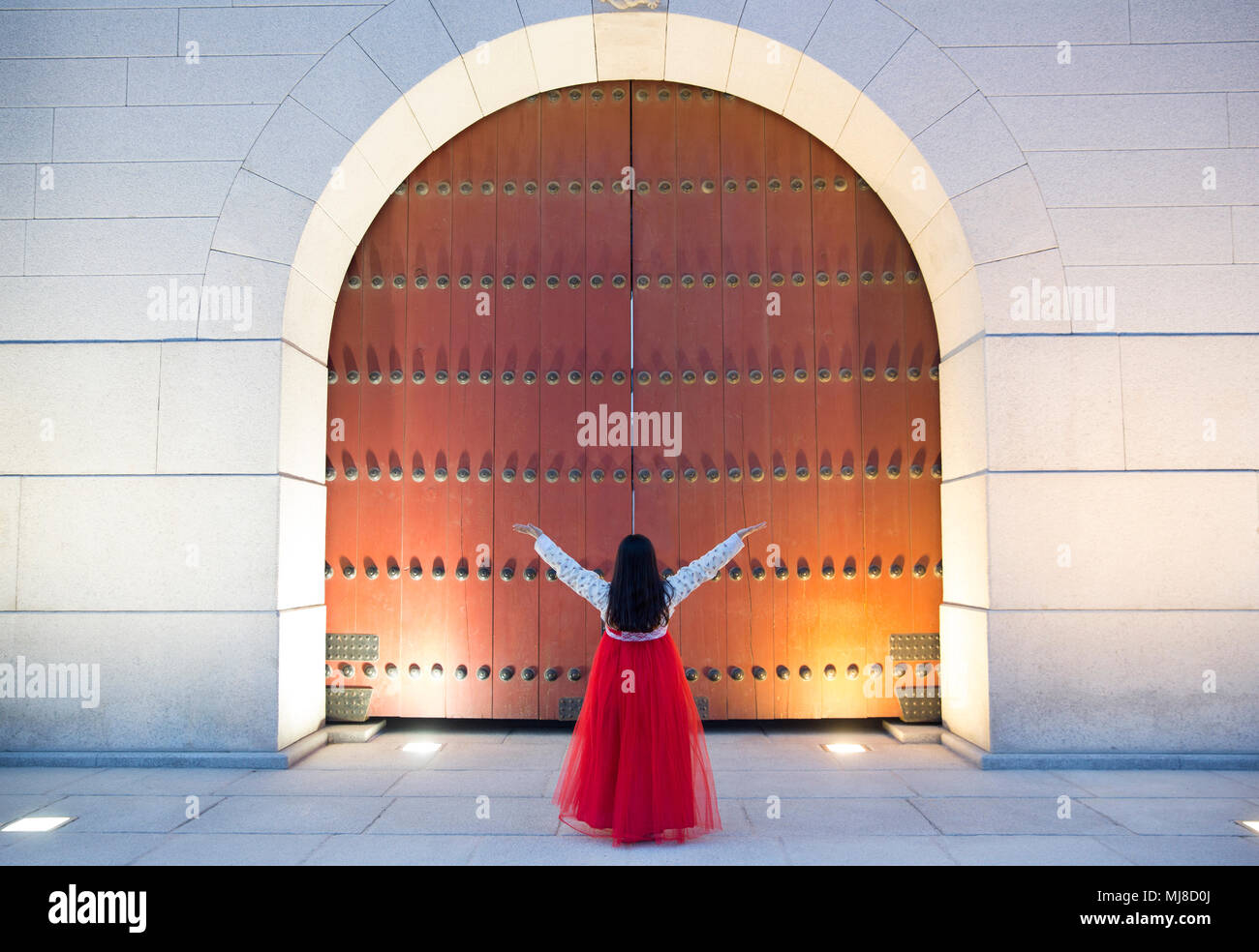 Rear view of woman wearing long jupe rouge debout avec son bras levés devant de grands porte en bois. Banque D'Images