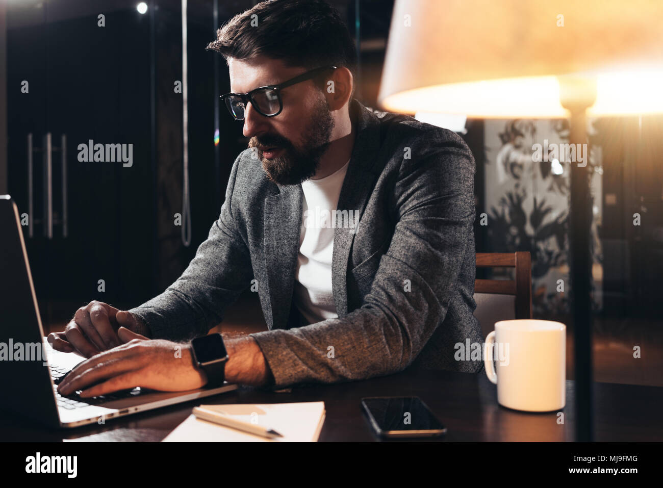 Jeune homme barbu travaillant dans l'espace loft dans la nuit. Un collègue est assis par la table en bois avec lampe et outils office. Directeur de projet à l'aide d'contempo Banque D'Images