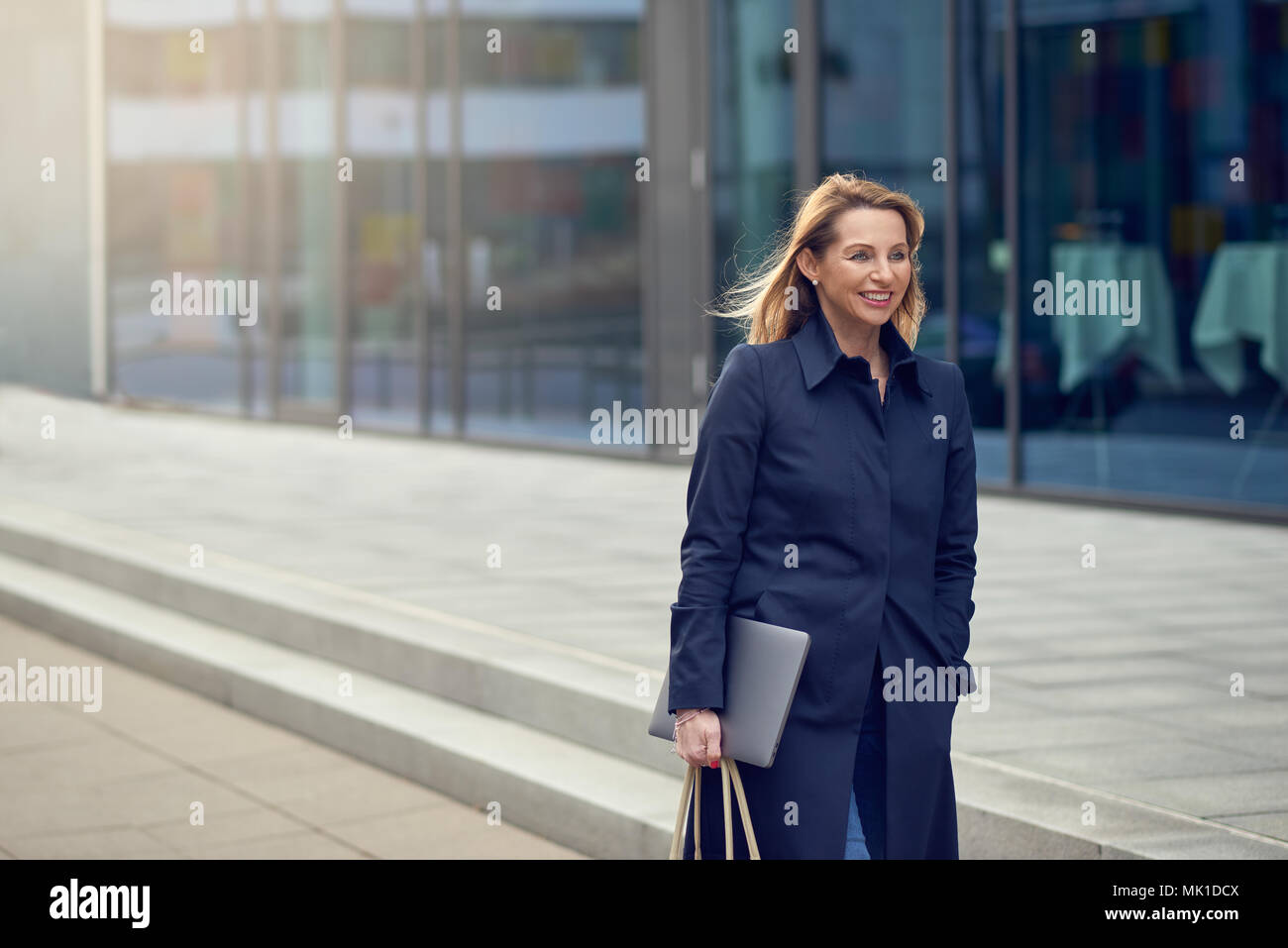Jolie femme blonde élégante de quitter son lieu de travail comme elle marche dans une rue urbaine high key calme sourire comme elle porte son portable with copy space Banque D'Images