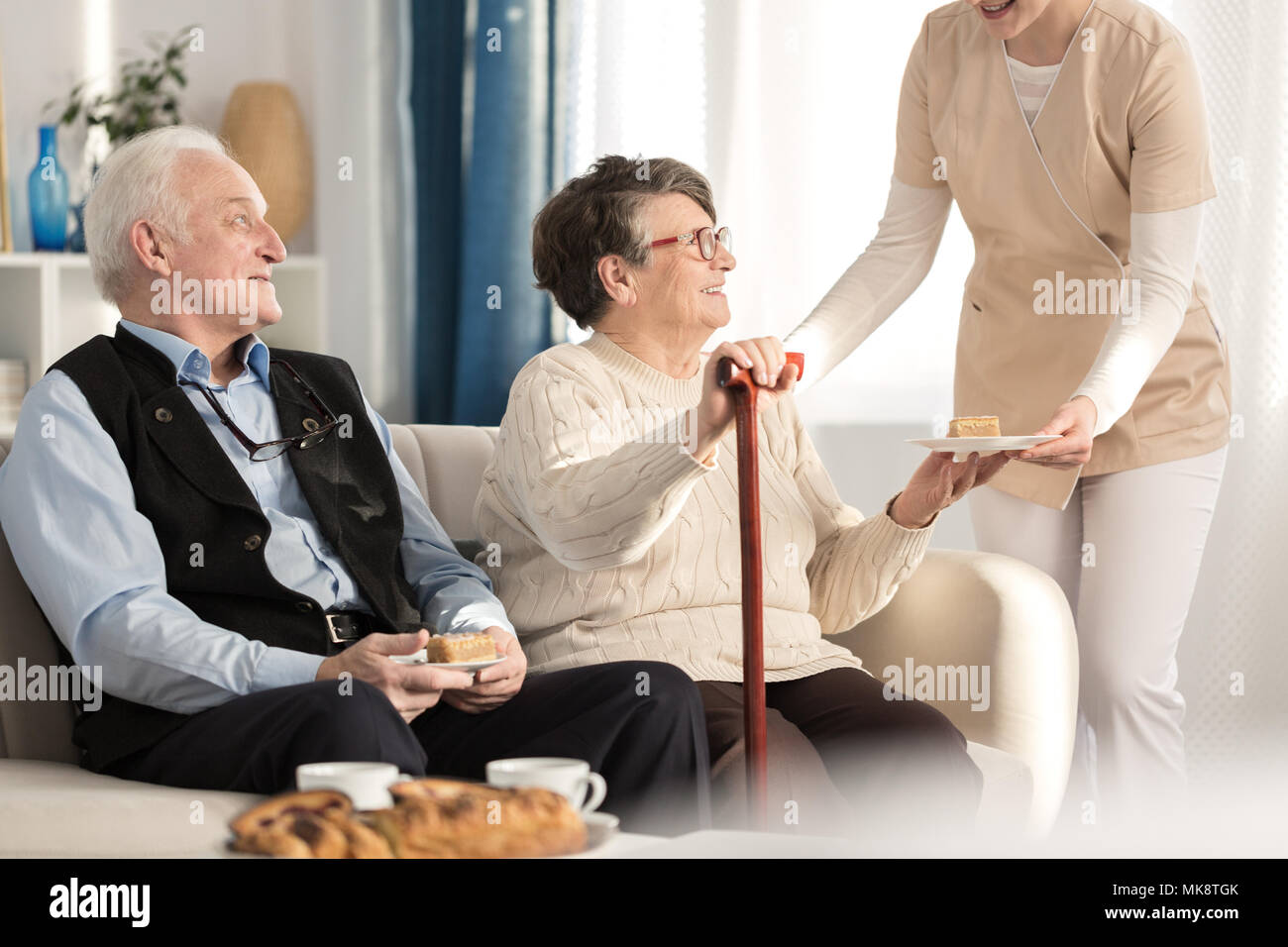 Couple gériatrique à l'arthrite assis sur une table et d'être servi un morceau de gâteau dans l'attente d'un rendez-vous chez le médecin lors d'une luxueuse salle de clini Banque D'Images