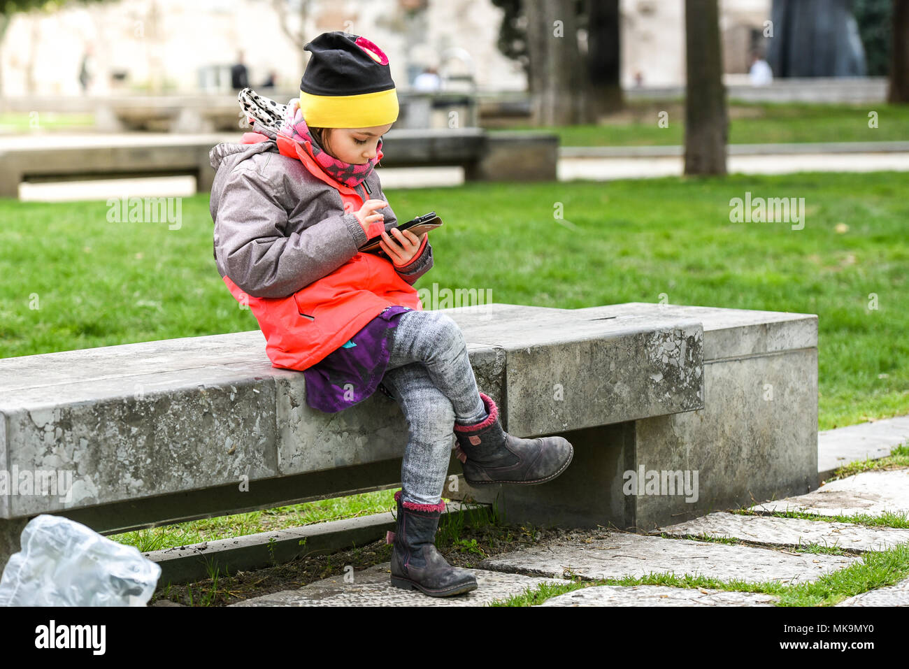 Peu de filles à l'aide d'un téléphone mobile en parc public. Au lieu de jouer à l'extérieur, l'enfant joue à des jeux sur téléphone, assis sur banc de granit. Chil Banque D'Images