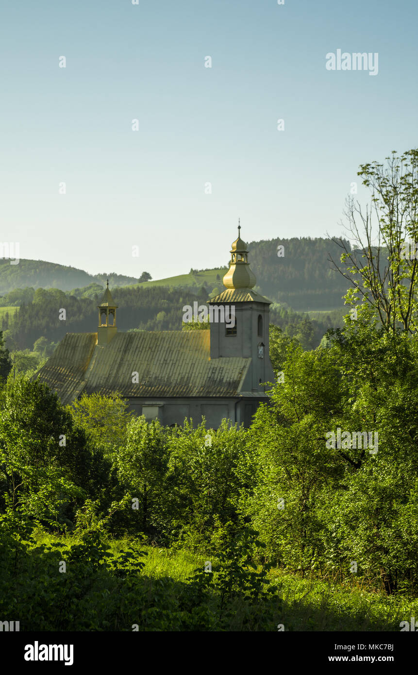 Église catholique avec tour d'or vert parmi les collines boisées au lever du soleil, dans Leszna Beskides de Silésie, Gorna, Pologne. Banque D'Images